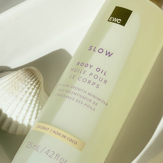 A bottle of EWC SLOW Coconut Body Oil resting on a white tray, with a seashell on the side.
