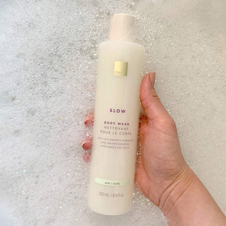 Hand holding a bottle of EWC Slow Aloe Body Wash against a bubble bath background