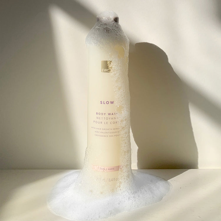 Bottle of EWC Slow Shea Body Wash with soap suds on a light background