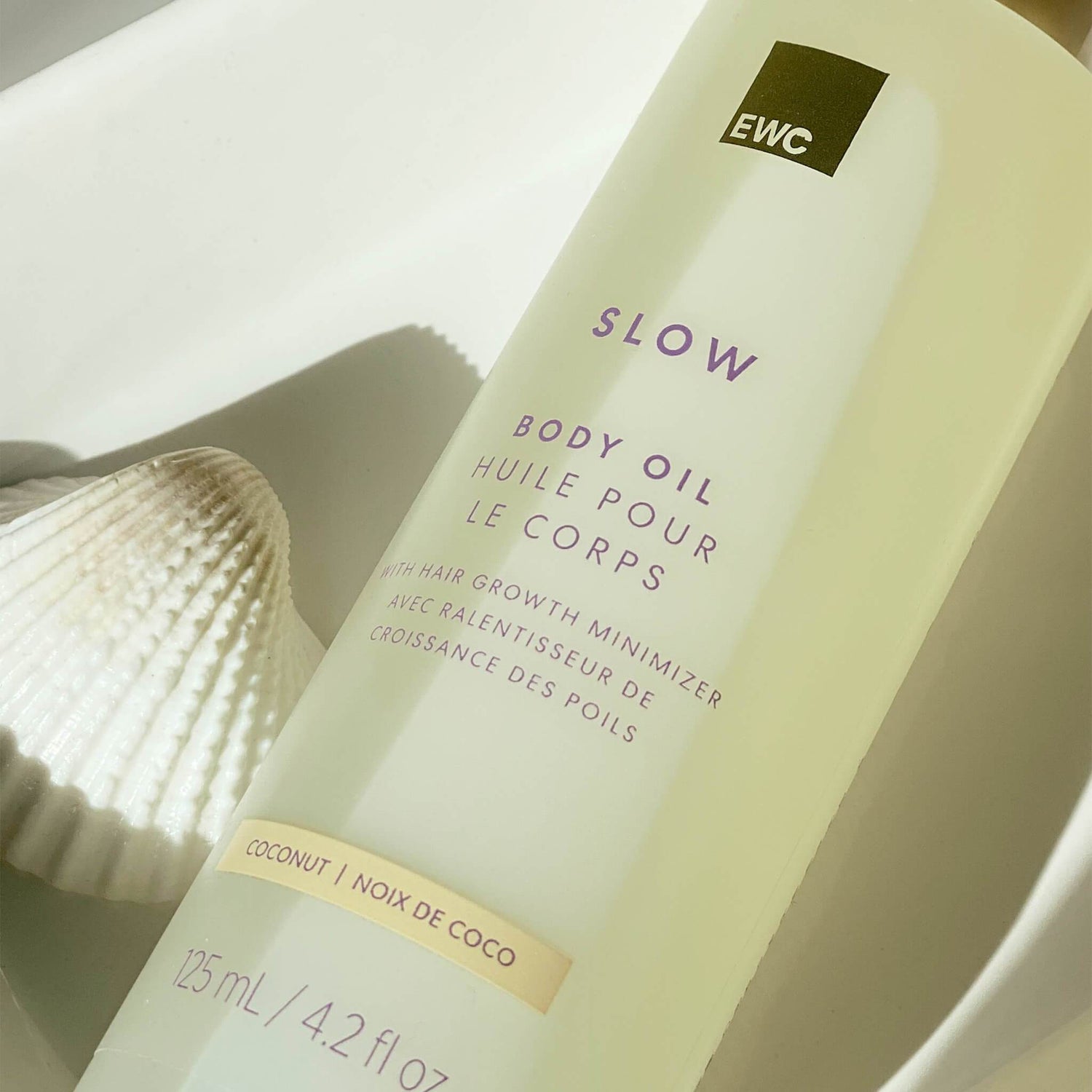 A bottle of EWC SLOW Coconut Body Oil resting on a white tray, with a seashell on the side.