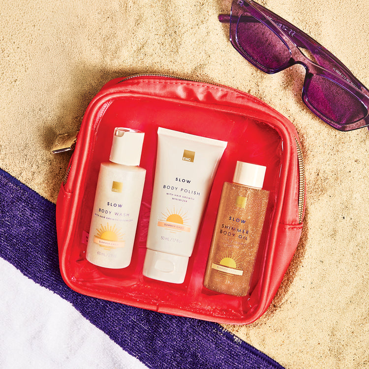 Travel-sized EWC SLOW® Summer Oasis collection, with products lined up inside the travel coral pouch. The kit is resting over beach sand and a purple and white towel, and a pair of purple sunglasses sits on the top right. 
